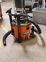 Front and side view of Ridgid orange shop vacuum with attached hose and two black nozzle accessories.