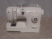 Front view of white Singer Inspiration sewing machine showing stitch selection dial, threading mechanism, and needle area.