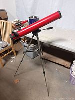 Red Tasco Model 33TR telescope mounted on aluminum tripod stand, set up in an indoor space near tables and storage bins.