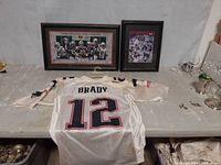 White Tom Brady official NFL jersey laid flat with back side showing number 12 and name BRADY, with two framed Super Bowl commemorative pictures behind.