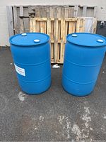Two blue plastic rain barrels standing upright outside on asphalt with wooden pallets behind them.