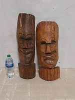 Both wooden tiki masks placed side by side with a water bottle for scale against a plain wall.