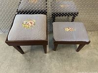 Two antique wooden stools side by side on a concrete floor with diamond plate metal background. Both stools have light grey fabric padded tops with hand-stitched floral needlework.