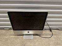 Front view of Apple iMac showing black screen, silver aluminum frame, Apple logo at stand base, and power cable.