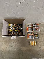 Box containing many loose used Duracell AA and AAA batteries, and five packs of Energizer batteries and one pack of GP alkaline batteries arranged beside the box on the floor.