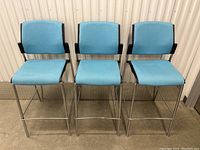 Front view of three blue bar stools with light blue fabric seats and backs, metal legs and footrests.