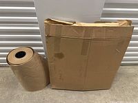 Large roll of kraft paper standing on the floor next to flattened cardboard sheets.