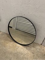 Round mirror with black frame leaning against a wall showing reflective surface and frame condition.