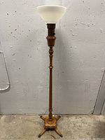 Full view of the wooden floor lamp standing against a concrete wall, showing the entire height and base with four legs ending in clawed metal feet.