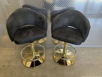 Pair of black upholstered official MGR casino chairs with curved backs, gold-tone adjustable height bases and circular footrests, on concrete floor with diamond metal plate wall background.