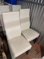 Four white leather Rozzini dining chairs with tall backs and wooden legs grouped together against a storage unit wall.