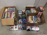 Cardboard boxes filled with VHS tapes and DVD cases showing titles like Lost, Sopranos, Seinfeld and various others spread in two boxes and some laid out in front.