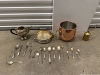 Full assortment of vintage cutlery and metalware arranged on the floor, includes spoons, forks, a teapot, a round dish, a large copper cup, and a small brass container.