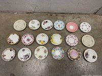 Top-down photo of 15 varied vintage tea saucers with floral patterns and different designs laid out on concrete floor.