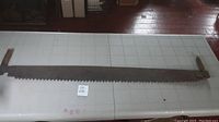 Full view of the antique two-man saw laid out on a table, showing the entire length of the saw with metal blade and wooden handle on each end.