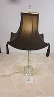 Table lamp with clear glass base on cream patterned tablecloth, black pagoda shade with tassels, lit from behind