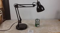 Black articulating desk lamp placed on fabric-covered surface next to a soda can to indicate size. Lamp is off.