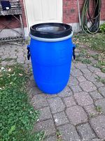 Blue plastic cylindrical barrel standing on a paved surface outside near grass, showing handles and black lid.