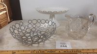 Metal basket fruit bowl, white metal dessert tray, and clear glass pitcher with floral embossing shown together on table.