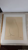 Full view of framed art print showing minimalistic black line drawing of a naked buttocks on beige background, and the white frame and matting.