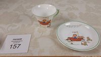 Cup and saucer set with child's illustrations and green rim on a tablecloth surface, accompanied by lot number card.