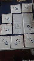Eight white cotton napkins and place mats folded, showing hand-stitched blue and red floral embroidery