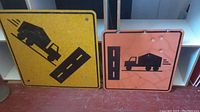 Two vintage metal road signs leaning against a wall, one yellow and one orange, showing their front graphics and overall condition.