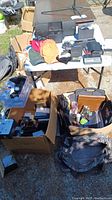 Overview photo showing the full lot with multiple boxes and bags containing electronics, hats, pens, and more item types on a white folding table outdoors.