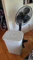 Honeywell pedestal fan and white plastic laundry hamper side by side on wood floor next to a wall.