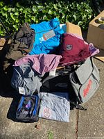 Full lot showing multiple jackets, shirts, and bags stacked on the ground outdoors with sunlight.