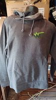 Gray hoodie on mannequin with drawstrings, front pocket, and green logo on chest.