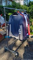 Full view of the metal rolling clothing rack with clothes on hangers (clothing not included) in outdoor setting.