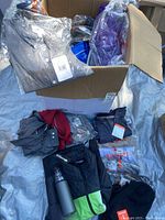 Lot with various folded clothing items inside and outside a brown cardboard box on a silver tarp, showing multiple sweaters and shirts in packaging.