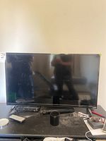 Full front view of a Vizio 40 inch LED TV on a table with remote, power cable, small black device and other small items around it.