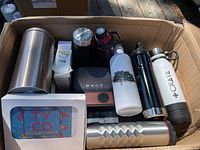 Wide shot showing all items inside a cardboard box, including bottles, speaker, and packaged swag.