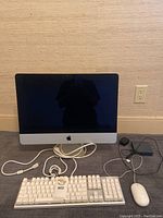 Front view of iMac display (powered off), showing white Azio keyboard with lot tag and white Apple mouse, along with power and data cables.