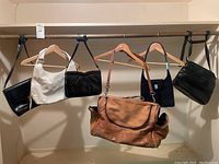 Six leather bags and purses hanging on a rail, including a brown leather duffle bag, a cream and black purse, and three black or navy purses.