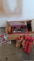 Boxed red Parmak electric fence charger, labeled Multi-Power Mark II, along with several red plastic fence posts and some wrapped accessories in packaging.