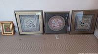 Four framed artworks arranged on floor including three square Oriental silk embroidered pieces and one smaller rectangular floral painting in gold frame.