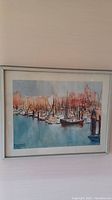 Framed watercolor painting showing a harbor with many boats, distant buildings in warm hues, and calm water in soft blue tones.