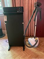 Full view of black Corby Windsor trouser press next to white and blue Steamfast steamer on parquet floor in room with pink curtains.