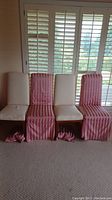Four chairs alternating between bare upholstery and pink striped slipcovers arranged in a row in front of window shutters