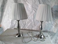 Photo showing two matching table lamps with stainless steel-like bases and white pleated shades on a table with a crumpled white background.