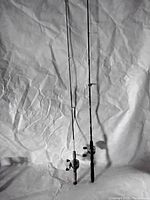 Two fishing poles side by side, shown vertically, against a white crumpled background. One rod has a cork handle, the other a black foam handle.