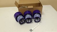 Three new Dyson vacuum filters with distinctive purple housings displayed in front of an open cardboard box. White cleaning brush included.