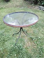 Full view of bistro table showing round mottled glass top, woven accent, and black metal legs on grass.
