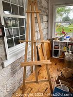 Wooden artist easel placed indoors near a window and shelving unit, showing the full height and adjustable components.