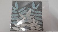 Paper pulp painting with leaf patterns in blue, white, and gray colors sealed in plastic.