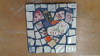 Top-down photo showing the full 6x6 inch square mosaic featuring floral and geometric crockery shards arranged in a heart motif with dark grout background.