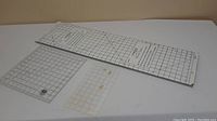 Three clear pattern cutting rulers and one long cutting board with measurement grids shown on tabletop